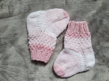 Zwei gestrickte Babysöckchen in Rosa und Weiß mit Rippbündchen auf grauem Stoff