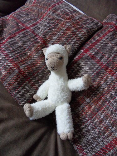 Gestricktes weißes Lamm-Amigurumi mit beigem Gesicht und Pfoten auf rot-brauner Decke