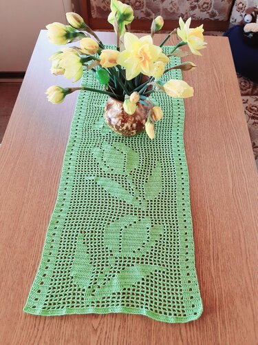 Grüner gehäkelter Tischläufer im Filetmuster mit Blumenmotiv auf Holztisch, braune Vase mit gelben Narzissen