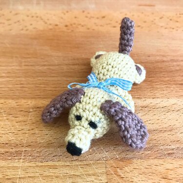beige gehäkelter Mini-Hund mit braunen Ohren und blauem Karoband auf Holz