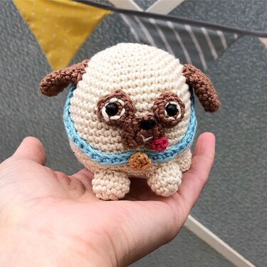 Hand hält einen kleinen gehäkelten Amigurumi-Hund in Creme und Braun mit blauem Halsband und Anhänger