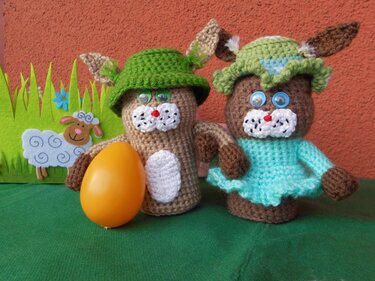 Zwei gehäkelte Hasen mit Hüten und Kleid neben orangefarbenem Ei und Filzschaf-Deko