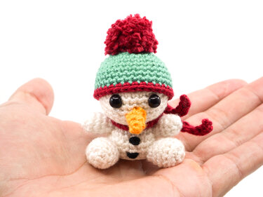 Palm-sized crocheted snowman amigurumi with green hat, red pom-pom and red scarf resting in an open hand