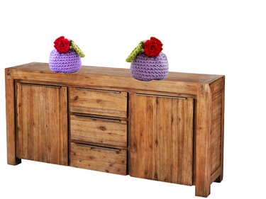 Zwei gehäkelte lavendelfarbene Dekoobjekte mit roten Blüten auf einer Holzkommode