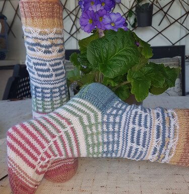 Zwei gestrickte Socken in Pastellfarben mit gestreiften und strukturierten Mustern; im Hintergrund eine Topfpflanze mit lila Blüten.