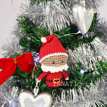 Gehäkelter Amigurumi-Weihnachtsmann mit roter Mütze und weißem Bart als Baumanhänger vor silbernem Lametta