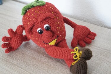 Handgehäkelte rote Erdbeerfigur (Amigurumi) mit Gesicht, Armen und braunen Schuhen auf hellem Holz