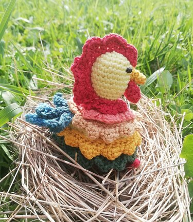 Mehrfarbig gehäkeltes Huhn mit rotem Kamm und blauen Schwanzfedern im Strohnest
