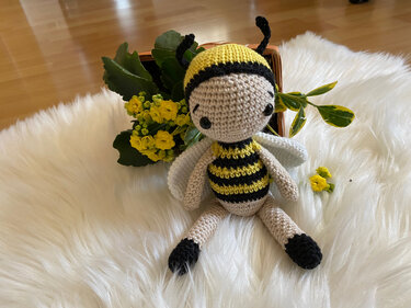 Gehäkelte Amigurumi-Biene in Gelb und Schwarz auf weißem flauschigem Teppich, mit gelben Blumen im Hintergrund