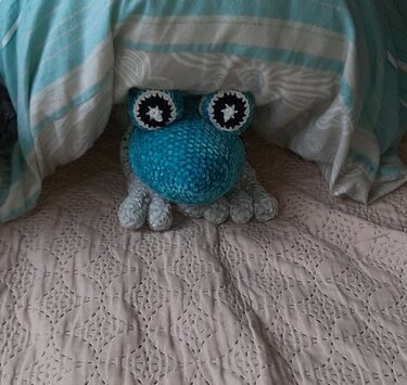 Verspieltes gehäkeltes Froschkissen in Blau und Türkis mit auffälligen Augen, auf einem Bett platziert