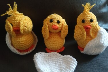 Drei gelbe Häkelküken mit schwarzen Augen und orangefarbenen Schnäbeln, zwei davon in weißen gehäkelten Eierschalen