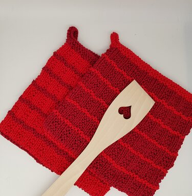 Two red and deep-red striped knitted potholders with hanging loops, shown with a light wooden spatula featuring a heart-shaped cutout.