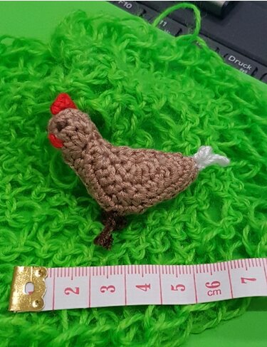 Mini gehäkeltes Amigurumi‑Huhn in Braun mit rotem Kamm auf grünem Garn, Maßband zeigt ca. 6 cm