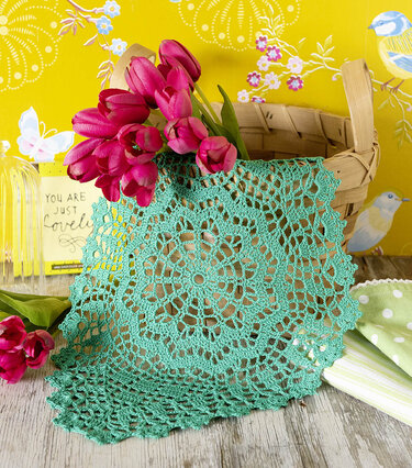 Türkises Häkeldeckchen mit floralem Spitzenmuster, drapiert an einem Holzkorb mit pinken Tulpen vor einer gelben Tapete mit Vögeln