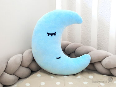 Plüsch‑Halbmondkissen mit aufgestickten Augen neben geflochtenem Nestchen im Babybett