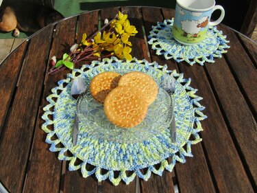 Zwei gehäkelte Deckchen in Blau und Grün auf Holztisch mit Teller, Keksen und Tasse