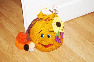 Handgehäkelter gelber Türstopper mit Gesicht, blauen Augen, Sonnenblume, lila Blüten, weißer Maus und kleinem Kürbis auf Holzfußboden