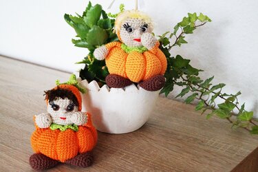 Zwei gehäkelte Amigurumi-Kürbisse mit Gesichtern, eine in einem weißen Blumentopf, die andere daneben auf Holz.