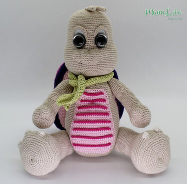 Gehäkelte Amigurumi-Schildkröte in Beige mit grünem Schal und rosa gestreiftem Bauch, sitzend vor hellem Hintergrund