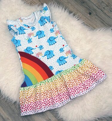 Weißes Kinderkleid mit blauem Elefantenmuster, seitlichem Regenbogen und buntem Herz-Rüschensaum, liegend auf hellem Teppich.
