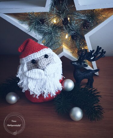 Gehäkelter Weihnachtsmann mit weißem Bart und roter Mütze neben Rentierfigur, Kugeln und beleuchtetem Stern