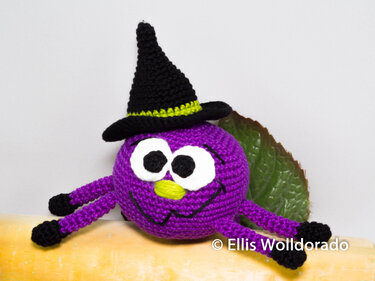 Lila gehäkeltes Amigurumi-Spinnenmonster mit schwarzem Hexenhut und grünem Band auf hellem Hintergrund.