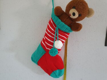 Gestrickter rot-türkiser Weihnachtsstrumpf mit weißen Streifen, Pompons und einem braunen Teddybär, der aus dem Strumpf schaut, an einer Wand hängend