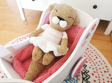 Gehäkeltes braunes Amigurumi-Bärchen im rosa-weißen Ballerina-Kleid in einer weißen Puppenwiege mit rosa Punkten.