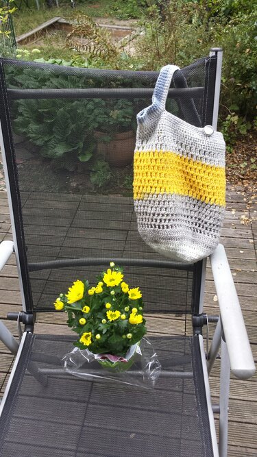 Handgehäkelte Henkeltasche grau und gelb mit Henkel und Knopf an einem Gartenstuhl, daneben gelbe Blumen
