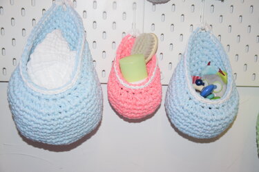 Drei gehäkelte Hängekörbe in Hellblau und Rosa an einer Lochwand, gefüllt mit Babyutensilien.