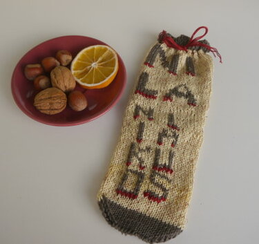 Beiger gestrickter Beutel mit roten Details und Kordel neben einer kleinen Schale mit Nüssen und einer Orangenscheibe