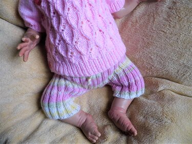 Rosa Babypullover mit Zopfmuster und gestreifte Strickhose auf hellbrauner Decke