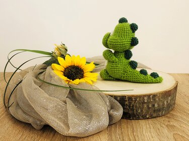 Grüner gehäkelter Amigurumi-Dinosaurier mit dunkelgrünen Rückenzacken, sitzend auf einer Holzscheibe neben einer Sonnenblume.