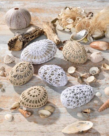 Gehäkelte Steine in Weiß und Beige, umgeben von Muscheln, Treibholz und einem Seeigel auf Holz