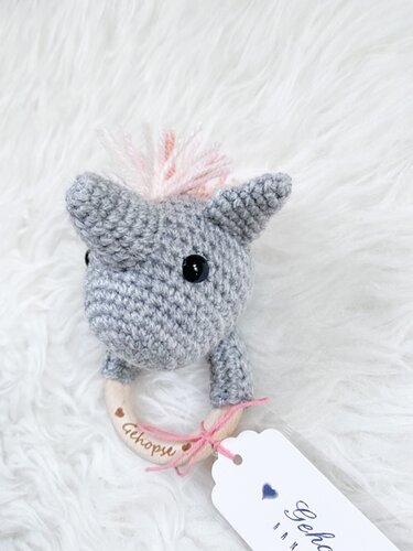 Gehäkelter grauer Einhorn-Greifling mit rosa Mähne und Holzring auf weißem Fell