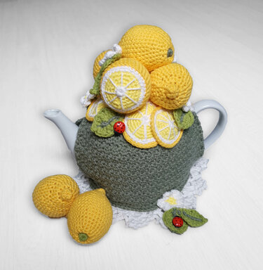White teapot covered by a green crochet cozy topped with yellow crocheted lemons, lemon slices, green leaves and small red ladybug accents, set on a white doily.