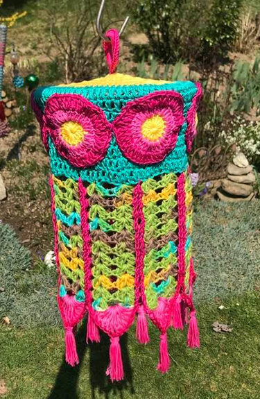 Gehäkelter Lampion in Türkis, Pink und Gelb mit großen Blumen und langen Fransen, hängend im Freien vor grünem Hintergrund.