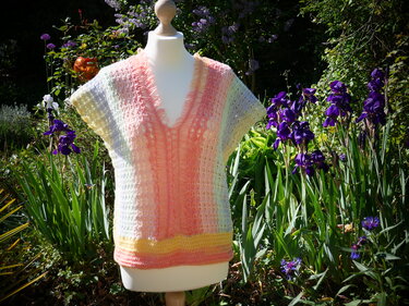 Gehäkeltes Pastell-Damen-Shirt mit V-Ausschnitt auf Schneiderpuppe im Garten bei lilanen Irisblüten.