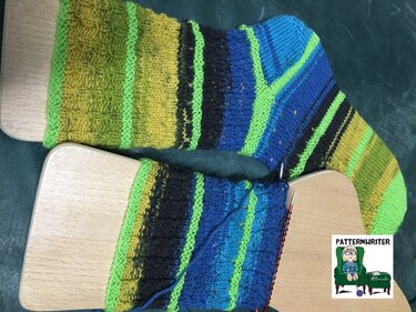 Zwei handgestrickte, gestreifte Socken in Grün, Gelb, Blau und Schwarz auf Holz-Sockenbrettchen, eine noch mit Stricknadeln.
