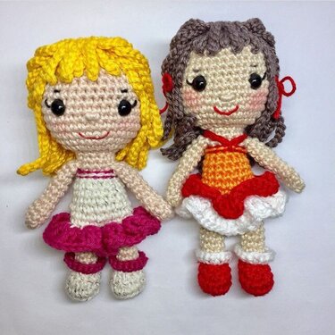 Zwei gehäkelte Amigurumi‑Mädchenpuppen, eine mit blonden Locken und pinkem Rock, die andere mit braunen Locken und rot‑orangefarbenem Kleid.