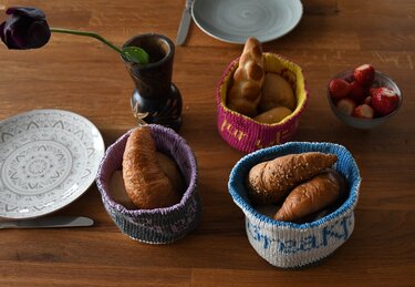 Drei gestrickte Körbchen in Blau-Weiß, Pink-Gelb und Grau-Lila mit Brötchen und Croissants auf einem Holztisch.