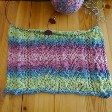 Gestricktes Lochmuster mit Farbverlauf (Grün, Blau, Rosa) auf Rundstricknadel, Wollknäuel daneben.