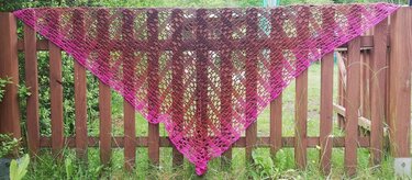 Gehäkeltes Dreieckstuch in Pink‑ und Brauntönen mit offenem Lochmuster, aufgehängt an einem Holzzaun