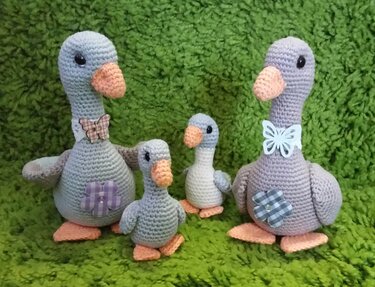 Vier gehäkelte Amigurumi-Gänse (zwei große, zwei kleine) mit orangefarbenen Schnäbeln und Stoffapplikationen auf grünem Hintergrund.