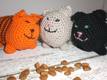 Drei gehäkelte Amigurumi-Katzen (orange, beige, schwarz) und Katzenfutter auf Holz.
