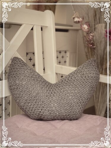 Graues V-förmiges Häkelkissen mit dichtem Strukturmuster auf hellviolettem Polster vor weißem Holzmöbel