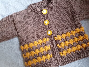 Braune gestrickte Babyjacke mit gelbem Muster und drei gelben Blumenknöpfen auf hellem Untergrund