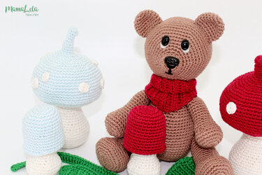 Gehäkelter brauner Teddybär mit rotem Schal, umgeben von roten und blauen Pilzen und grünen Blättern vor hellem Hintergrund.