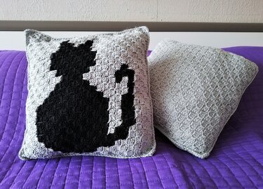 Zwei gehäkelte Kissen auf einer lila Decke, eines mit schwarzer Katzensilhouette.