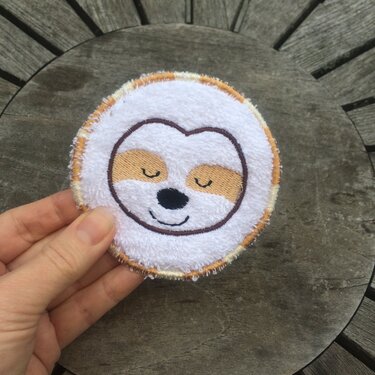 Hand hält rundes weißes Stickpad mit gesticktem Faultiergesicht auf Holzuntergrund.